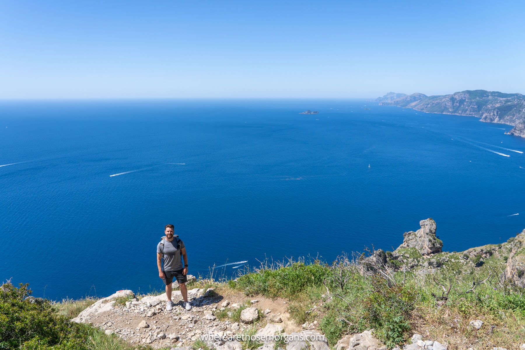 Photo of Mark Morgan in hiking gear standing on a stone path on a cliff edge with far reaching views over a deep blue colored sea behind on a clear day in the Amalfi Coast