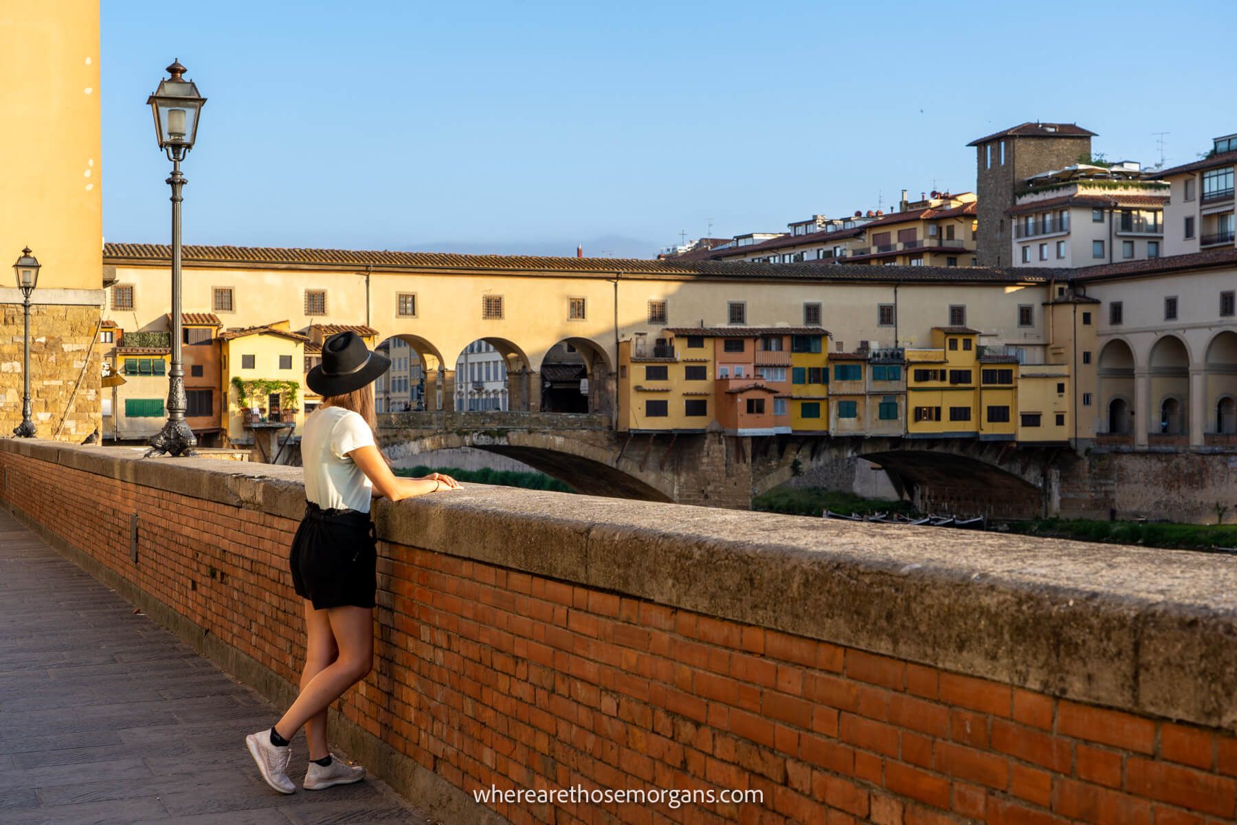 Photo of Kristen Morgan wearing a hat and stood next to a wall looking over it towards a river with a bridge on a sunny day in Florence