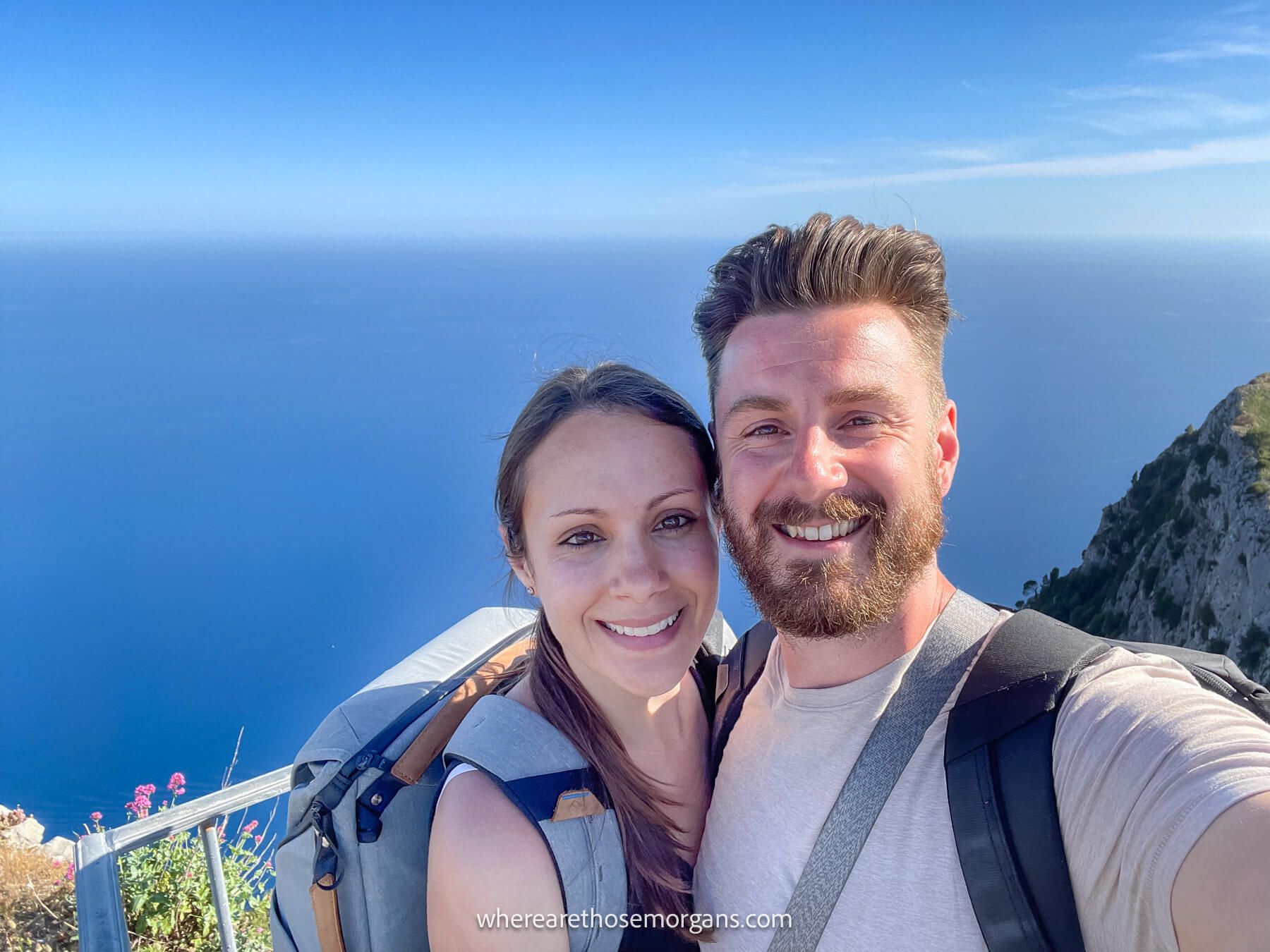 Photo of Mark and Kristen Morgan from Where Are Those Morgans taking a selfie from an elevated viewpoint overlooking the ocean on a sunny day in Italy