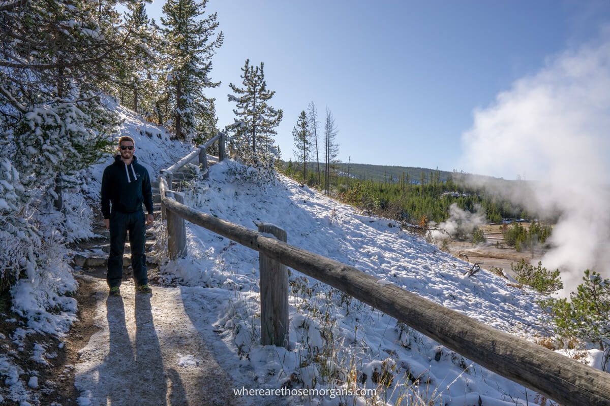 Hiker in winter clothes walking on curving a path with steps and a wooden barrier next to trees with lots of snow on the ground and sun in the sky