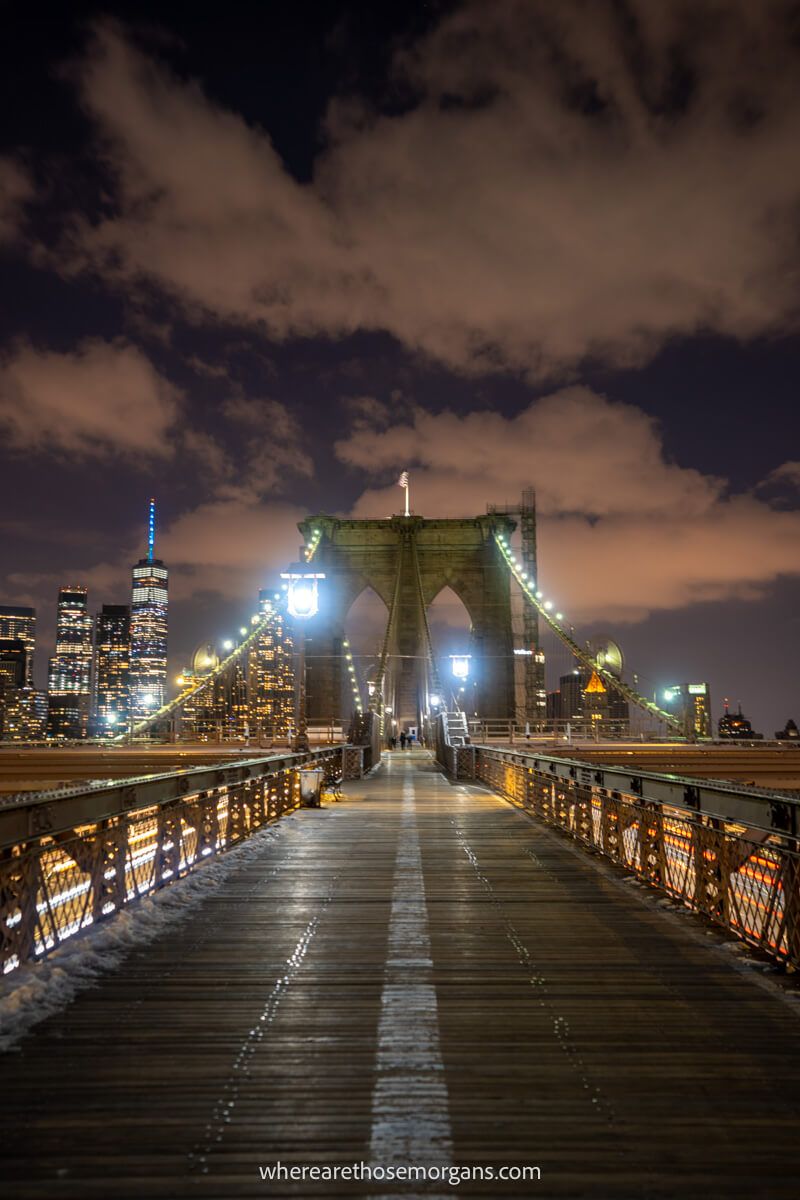 Photo of a narrow pedestrian walkway on the Brooklyn Bridge leading to Lower Manhattan at night with clouds in the sky