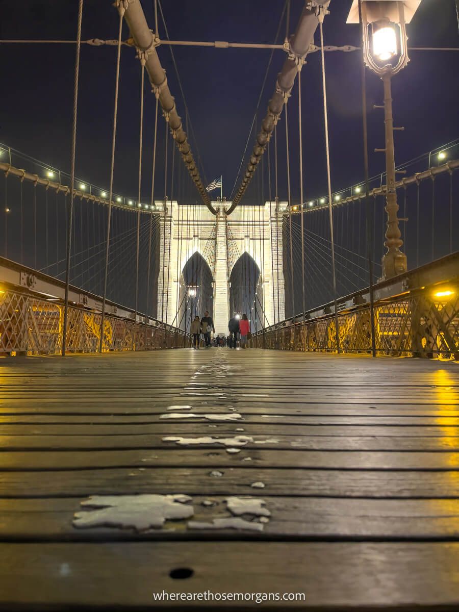 Photo of a long and narrow wooden boardwalk leading to a buttressed double arch at night with a street light