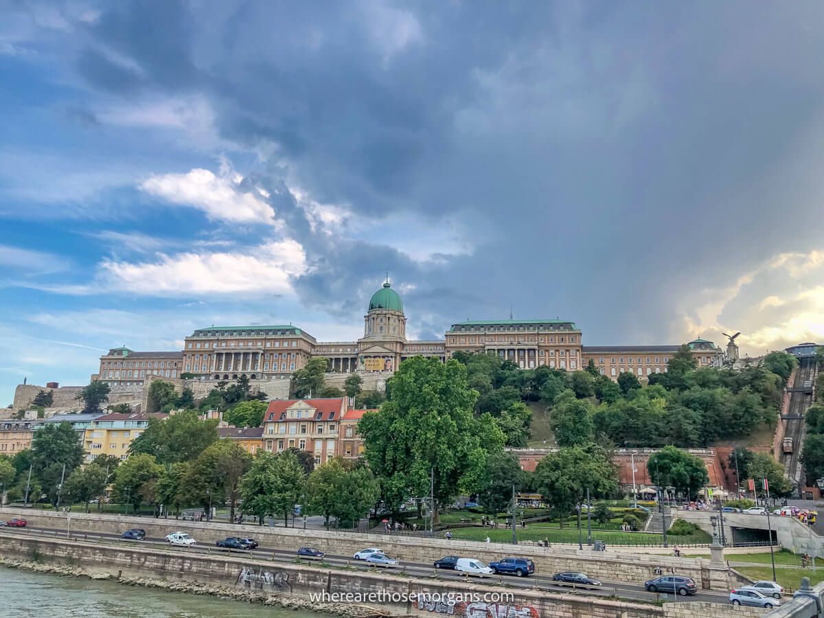 Photo of an old castle on a hill in Budapest with trees and a river in front on a cloudy evening