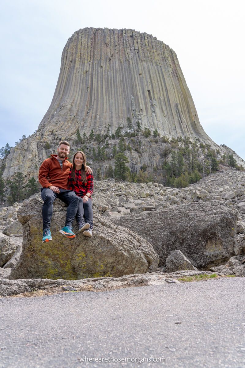 Photo of a couple sat on a boulder with the massive Devils Tower rock formation bursting into the sky behind
