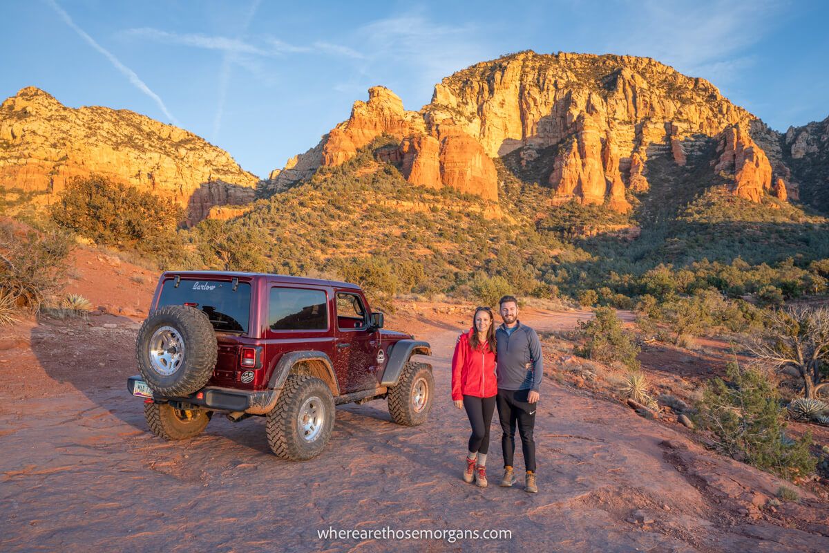 Couple standing together next to a maroon colored jeep on red rocks with rocky spires in the background at sunset