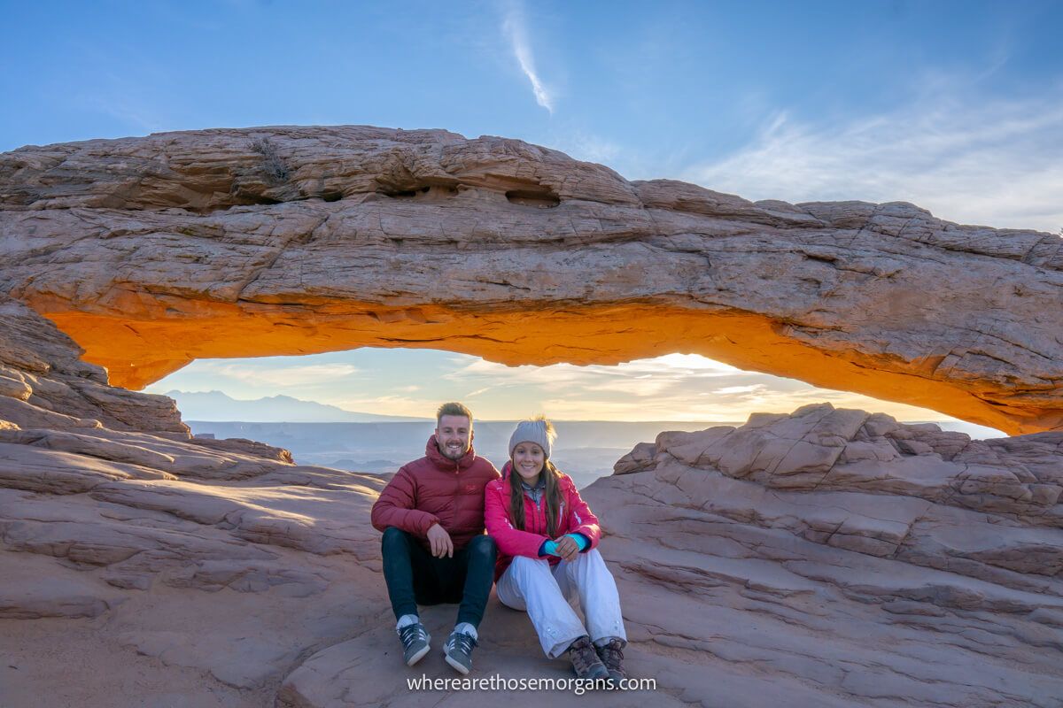 Couple sat next to each other on a rocky shelf with a natural window shaped rock formation behind
