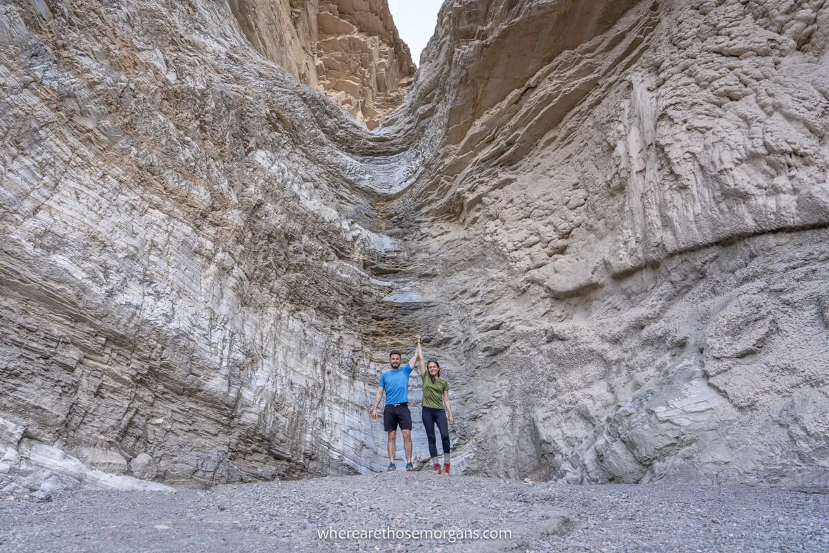 Couple hiking together and holding each others arms in the air at the bottom of an eroded waterfall in a dry canyon