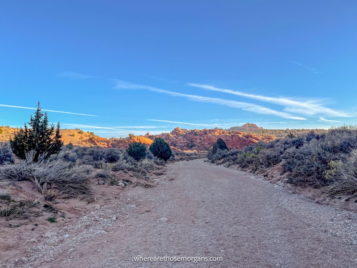 Photo of a dry river bed flanked by desert vegetation and leading to red rock formations on a sunny day with blue sky