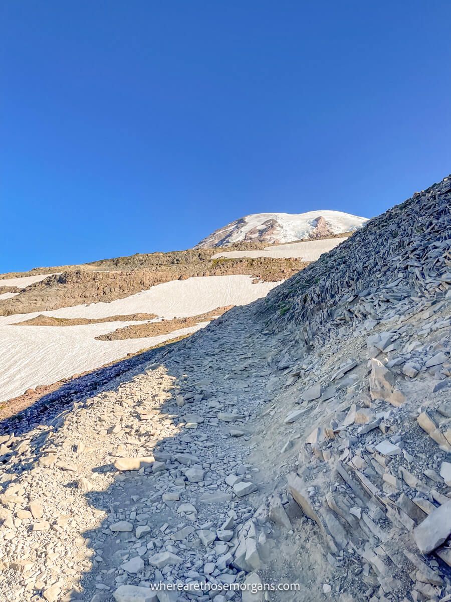 Photo of dry, rocky and dusty path leading to a mountain summit with snow on the ground under a blue sky