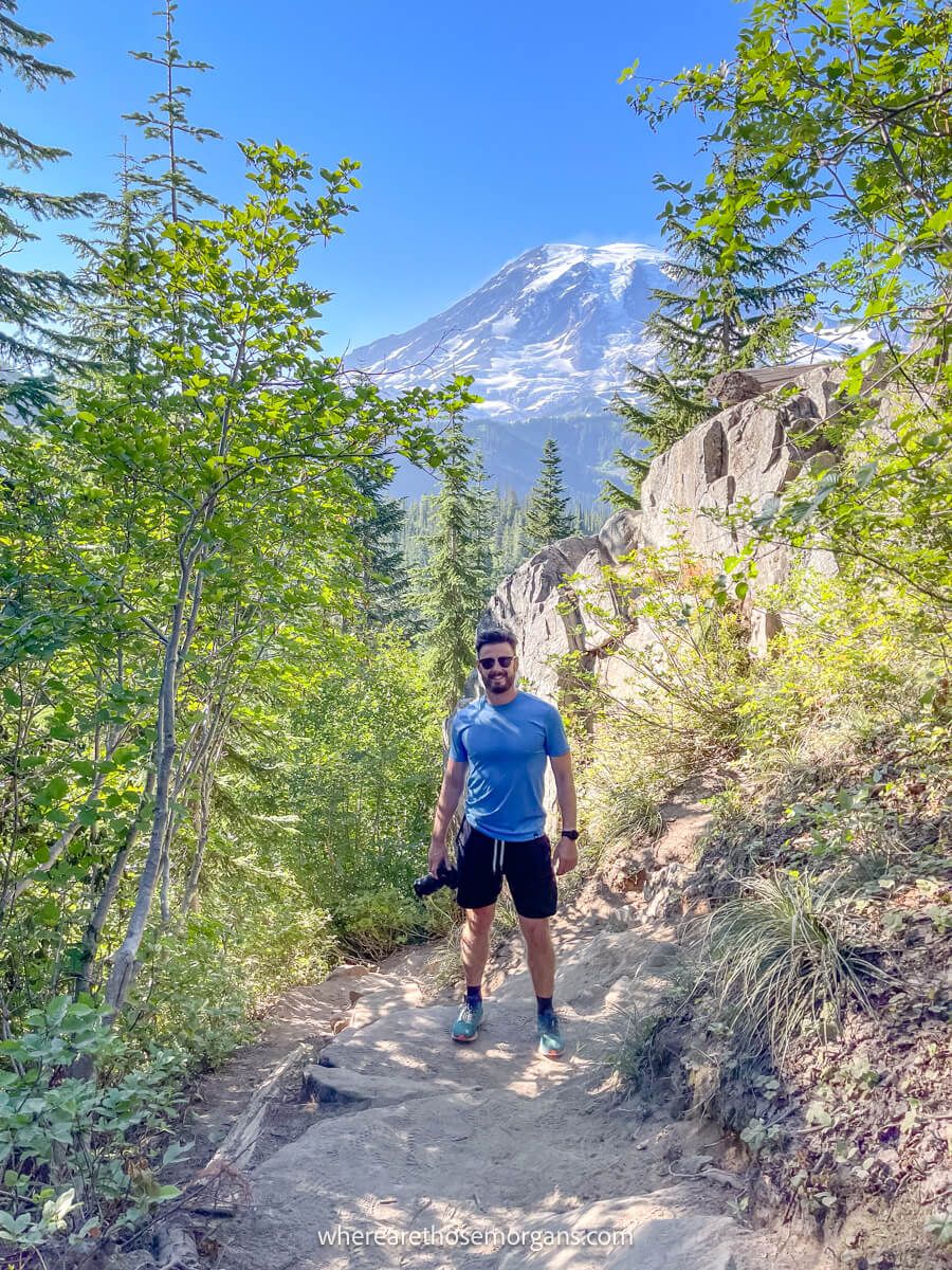 Photo of Mark Morgan from Where Are Those Morgans hiking in a blue t-shirt and shorts holding a camera on a dirt path with views over Mt Rainier behind on the Bench and Snow Lakes Trail