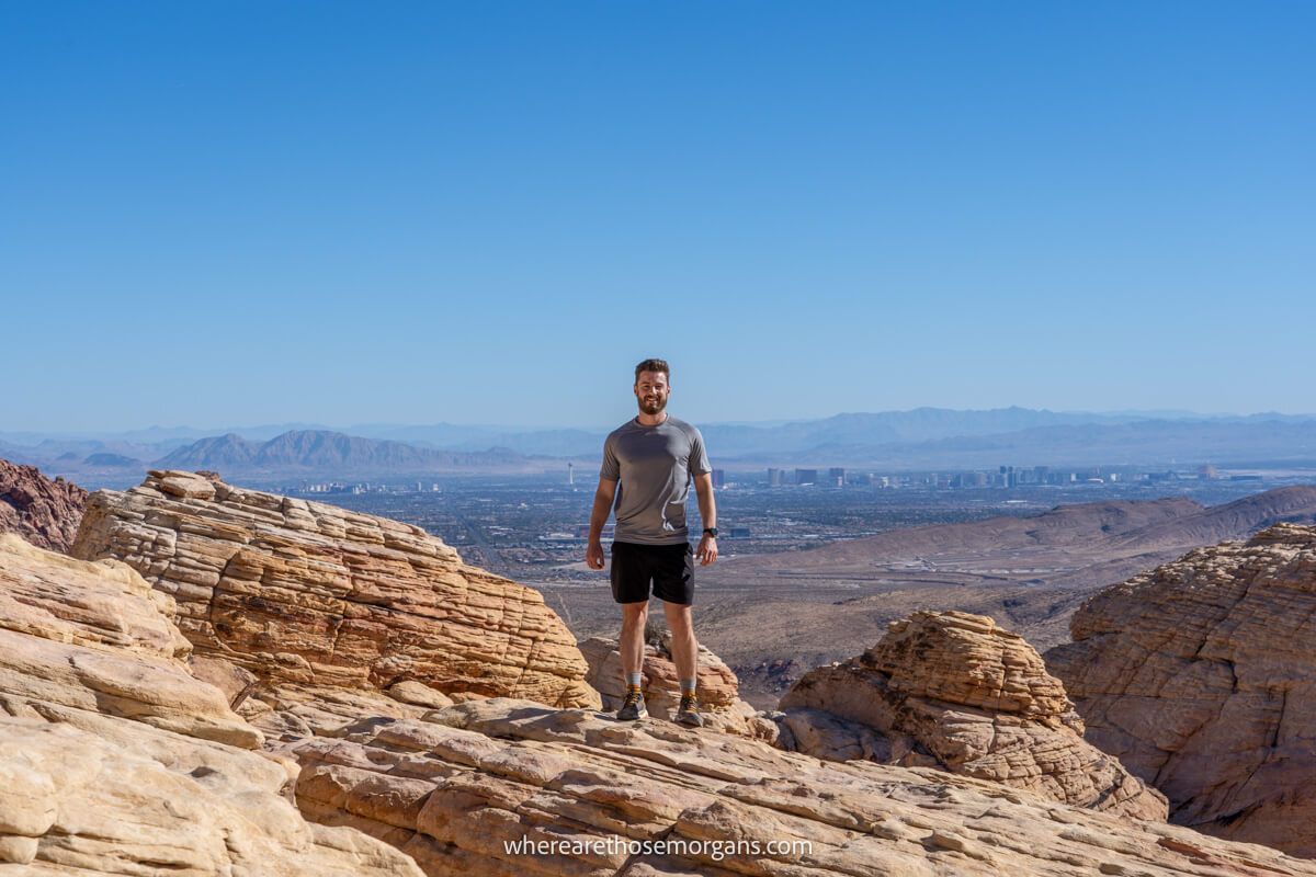 Hiker stood alone on a rocky summit with far reaching views over a city in the distance and a clear blue sky