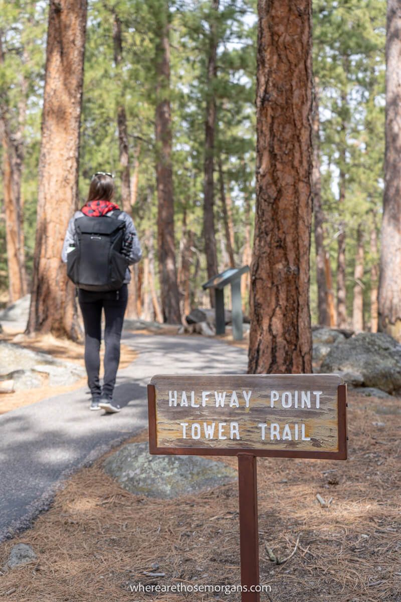 Photo of a hiker walking on a paved path through a light forest with a wooden sign marking the half way point of the trail