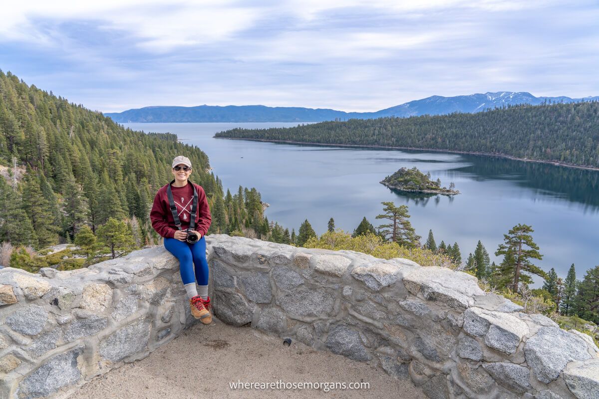 Photographer with blue pants and maroon sweater sat on a wall with camera in hand, and a lake with trees and thick clouds behind
