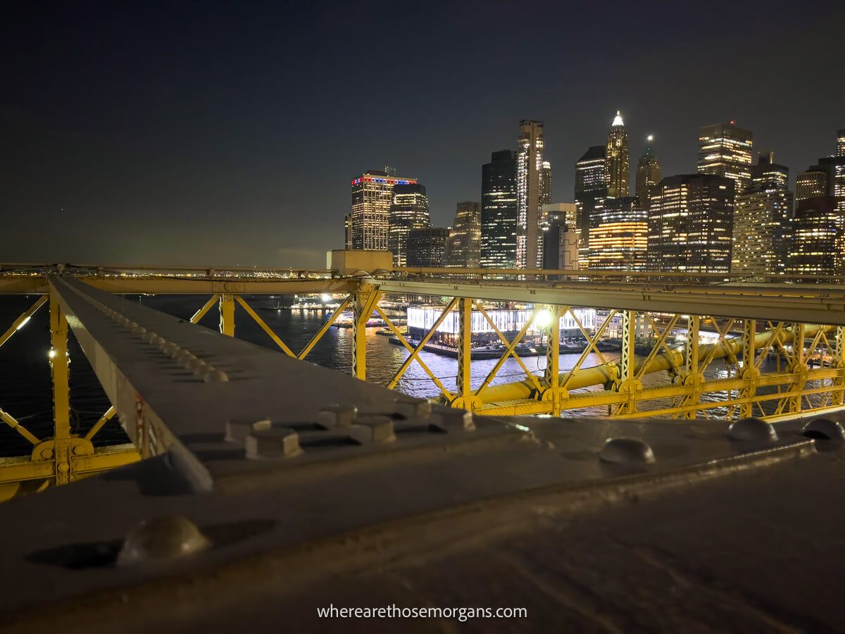 Photo of Lower Manhattan skyline lit up at night taken from the Brooklyn Bridge with steel girders above a lane of traffic in the foreground