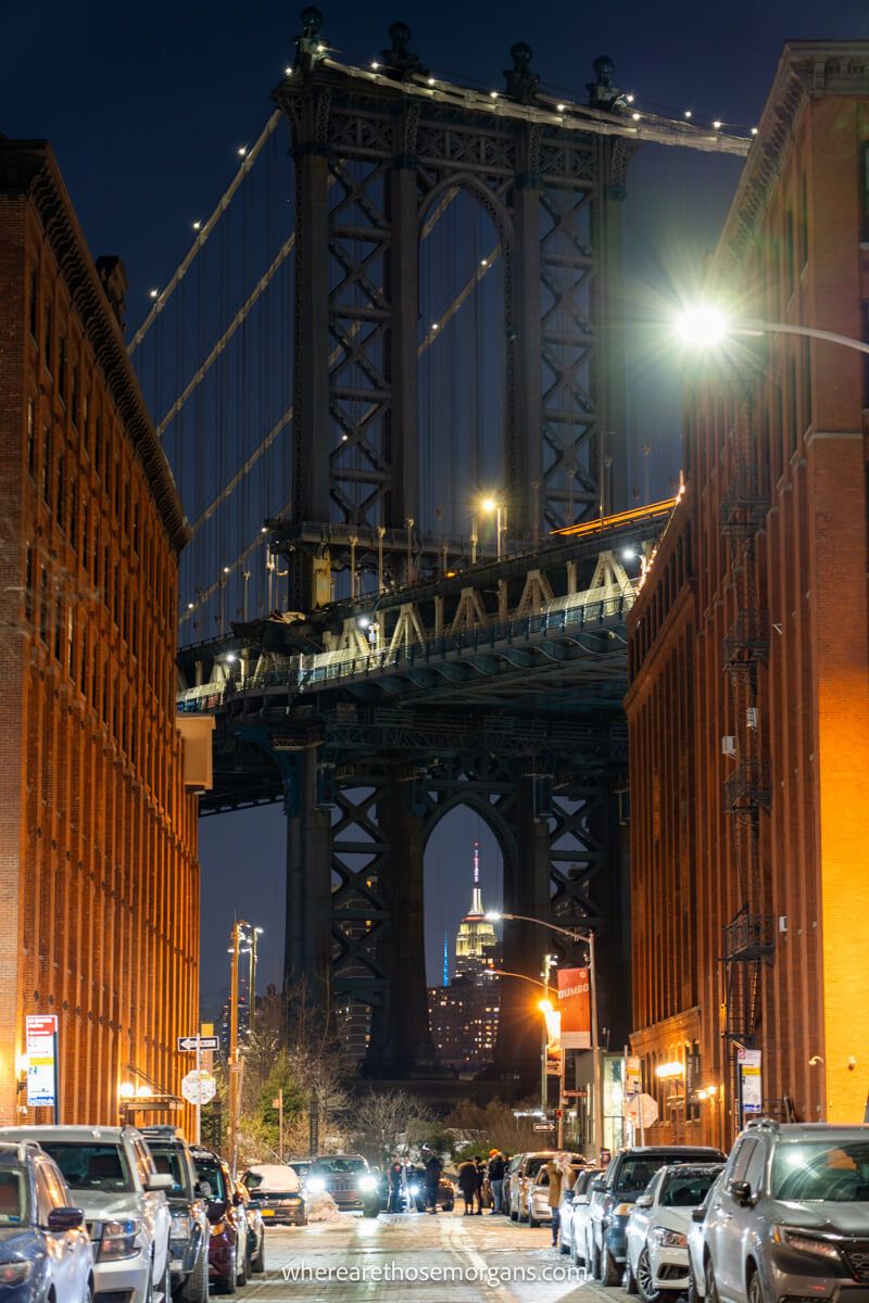 Photo of a street with tall brick buildings and cars parked leading to a massive bridge at night