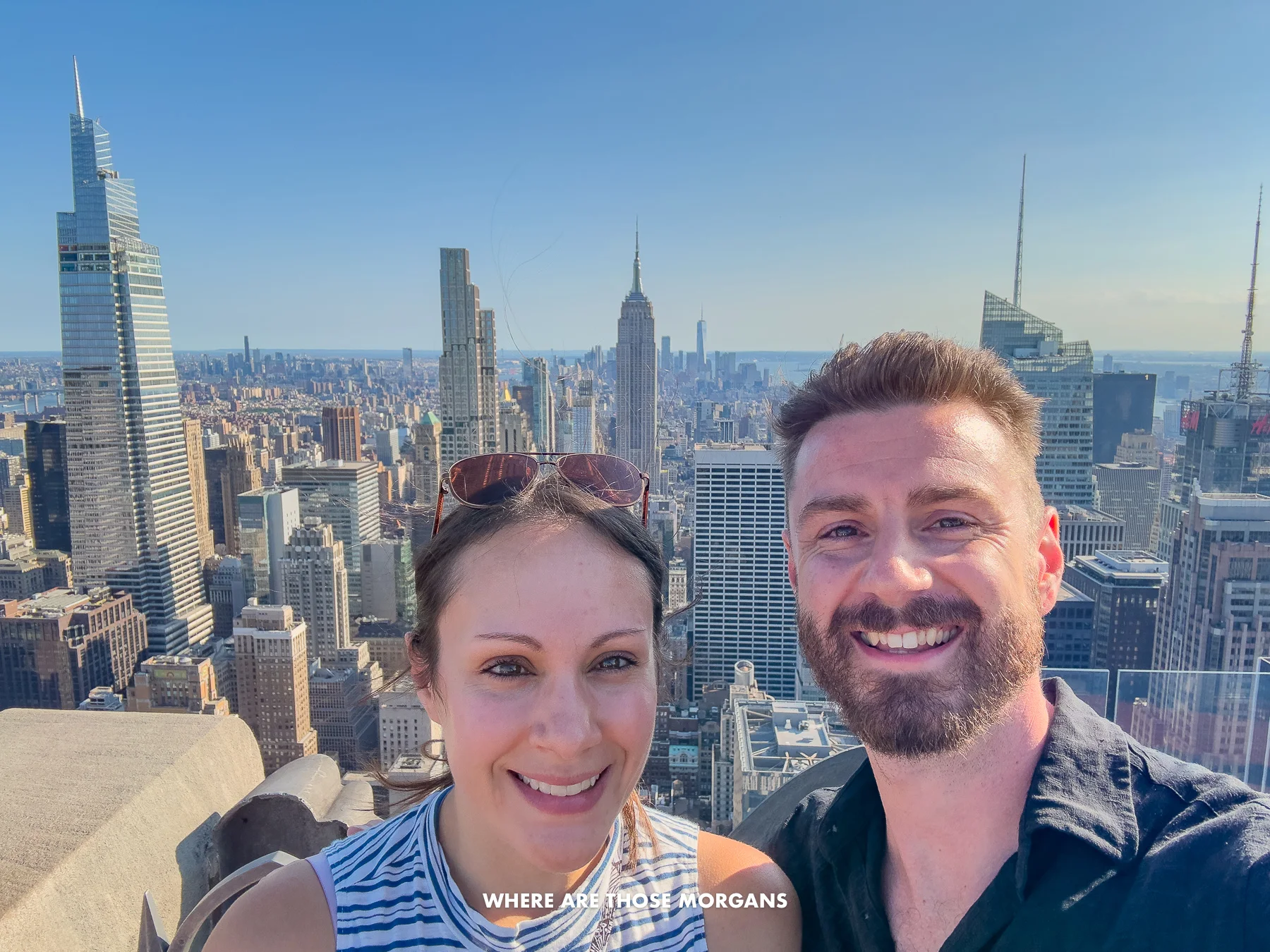 Mark and Kristen taking a selfie on the open top floor at Top of the Rock on a sunny and clear day with views over NYC behind