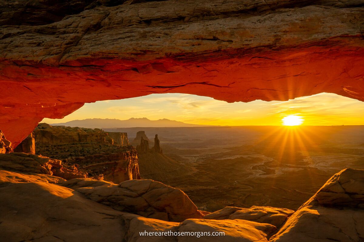 Photo of a striking sunrise on a distant horizon behind a wide open and deep valley, as well as an eye shaped natural window formed at Mesa Arch in Canyonlands National Park