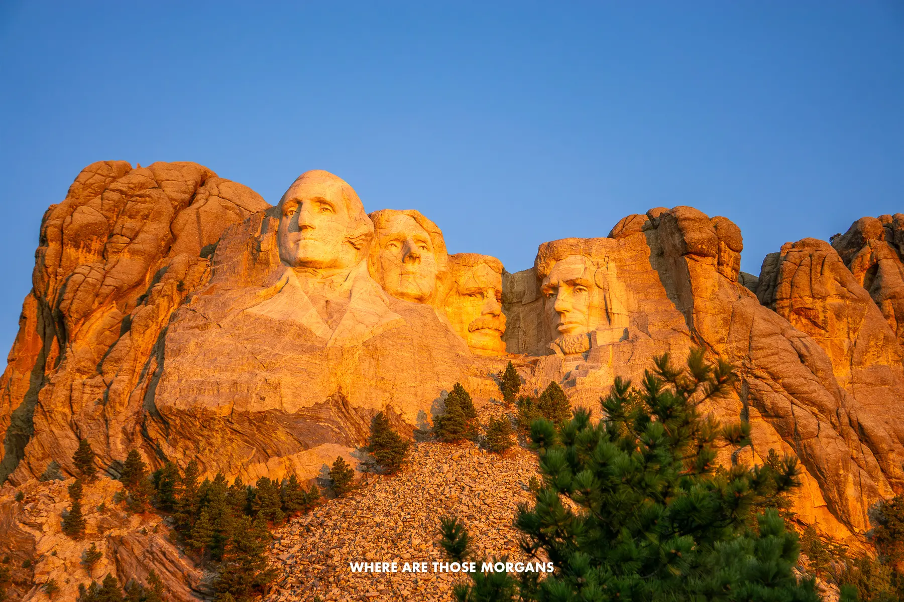 Looking up at Mt Rushmore carved into the mountain at sunrise as it glows orange against a clear deep blue sky