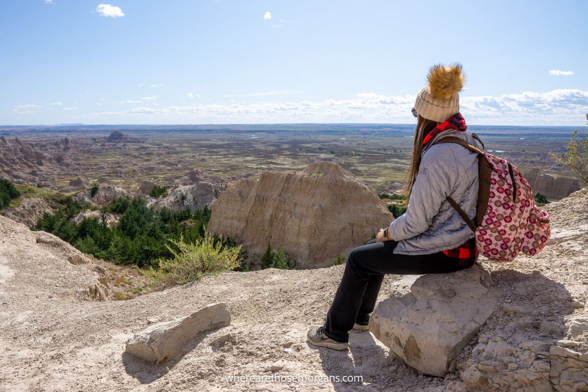 Hiker with backpack and hat sat on a small rock overlooking a far reaching view of rocks, trees and plains on a clear sunny day