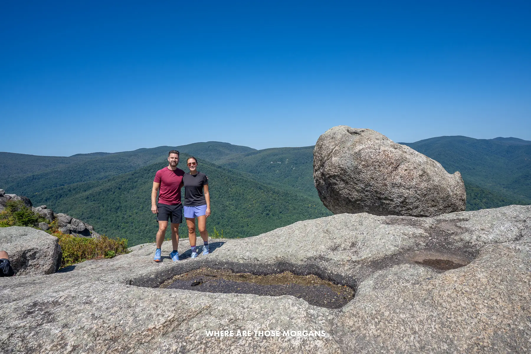Mark and Kristen from Where Are Those Morgans standing on rocks next to a big boulder with views of rolling hills behind at the summit of Old Rag Trail