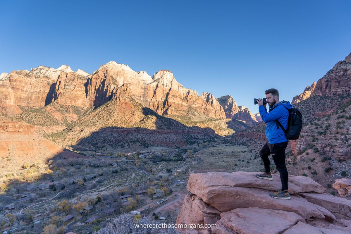Photo of a hiker with a camera at a rocky summit looking down over Zion visitor center and the Pa'rus Trail running through the valley floor