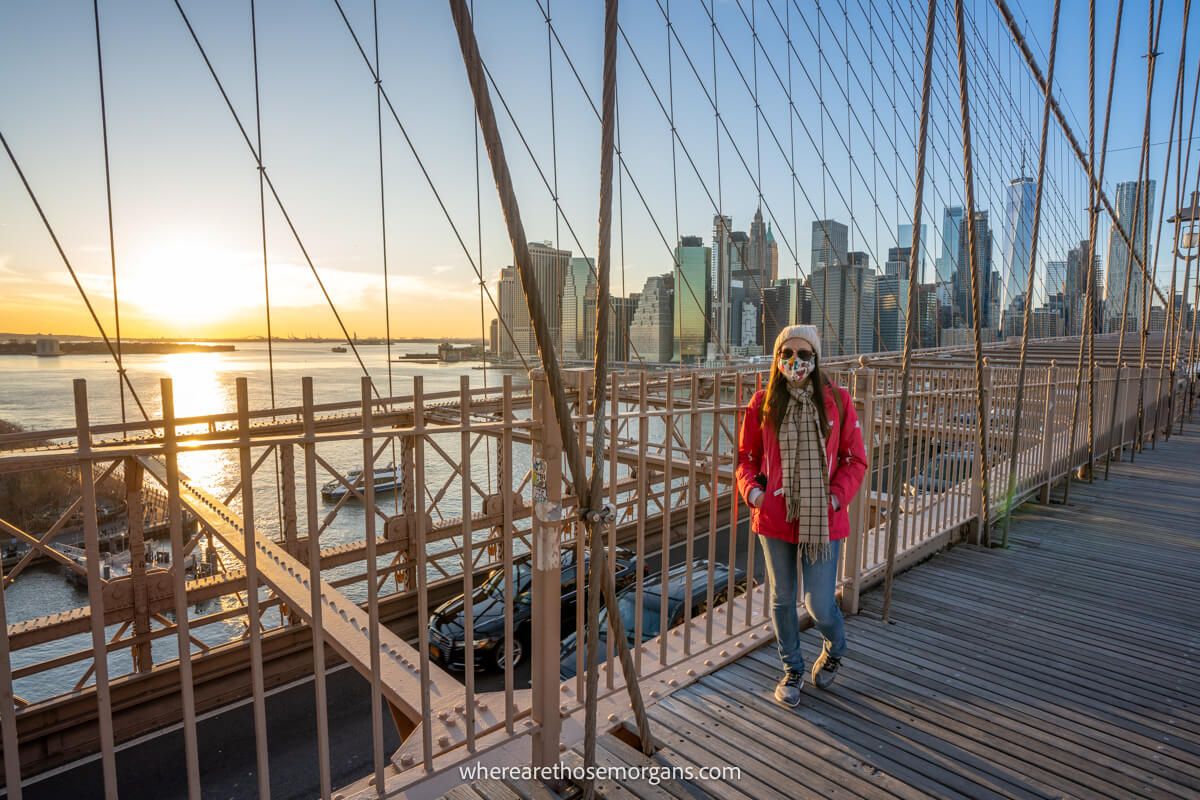 Photo of a tourist wearing coat, hat and mask on the Brooklyn Bridge pedestrian walkway with the sunset behind on the East River with Lower Manhattan skyline on the other side