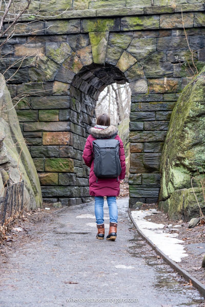 Photo of a tourist wearing coat and backpack walking underneath a small stone arch in New York