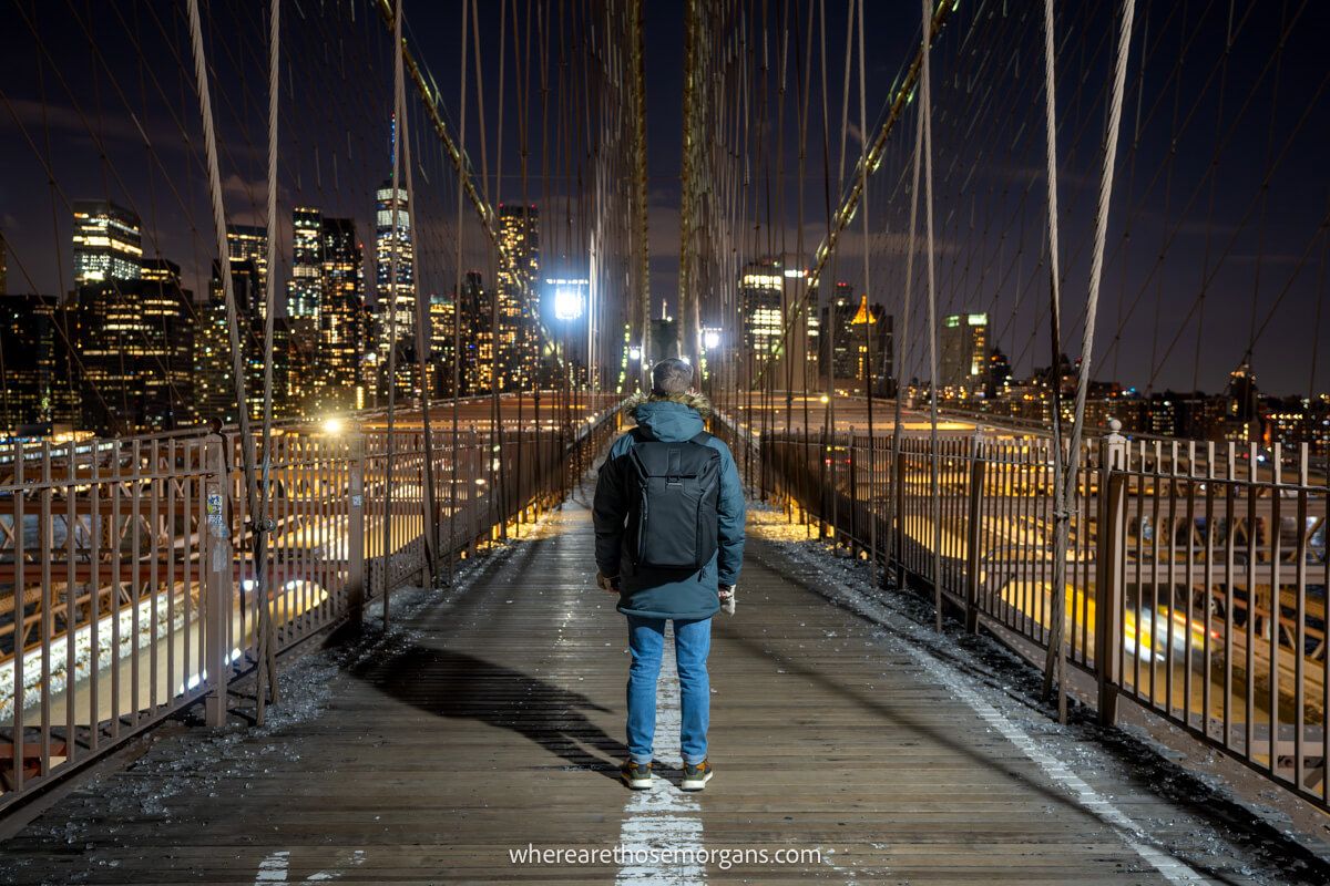 Photo of a tourist wearing winter coat and gloves walking the Brooklyn Bridge at night