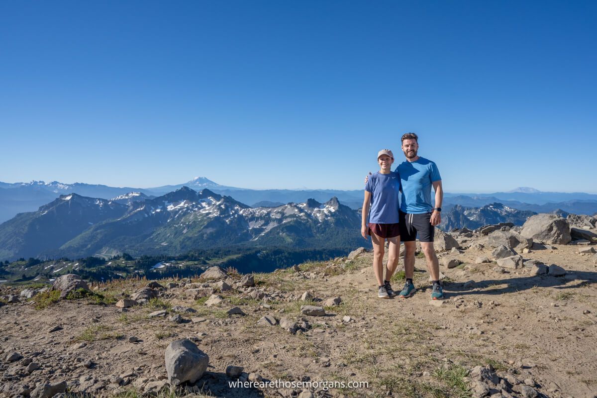 Photo of Mark and Kristen Morgan from Where Are Those Morgans standing together on a rocky summit with far reaching mountain views behind under a clear blue sky at the top of Skyline Trail hike in Mt Rainier