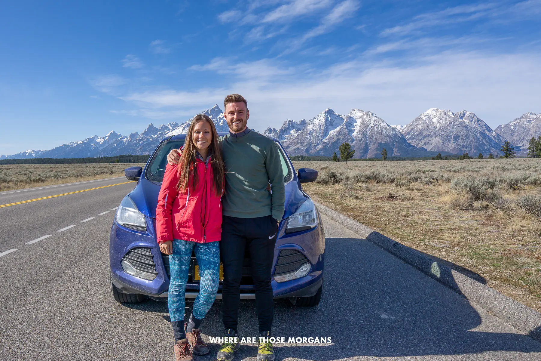 Mark and Kristen from Where Are Those Morgans standing together in front of a blue car on an empty road with prairie and mountains behind on a USA road trip