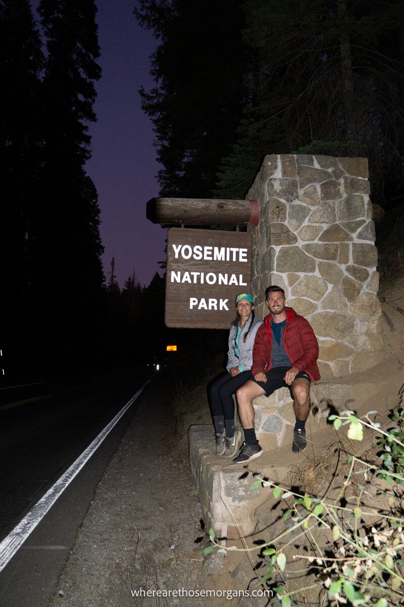 Couple sat together on a brick wall next to a road at night with a Yosemite National Park sign